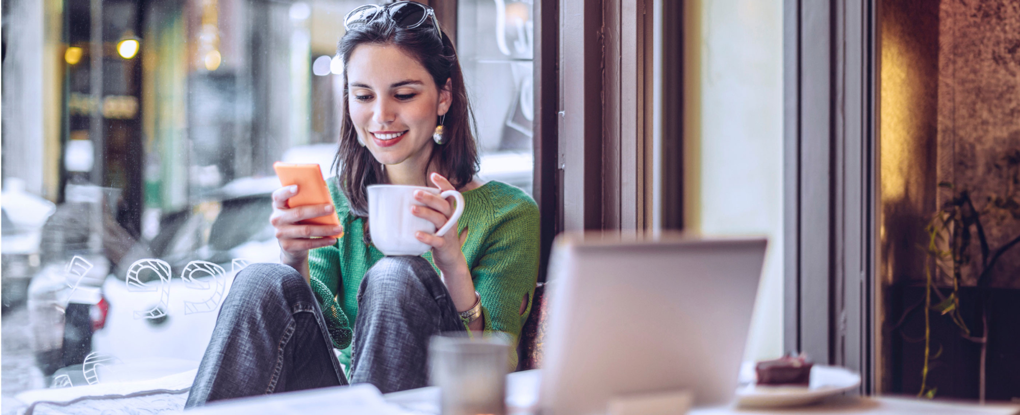 A young woman sat inside a cafe by a window smiling and looking at her phone with a cup in her hand and a laptop on the table next to her
