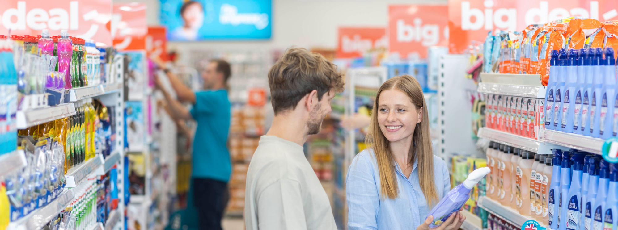 A man and a woman standing in the isle of a supermarket and smiling, in the background a shop assistant stacks some shelves