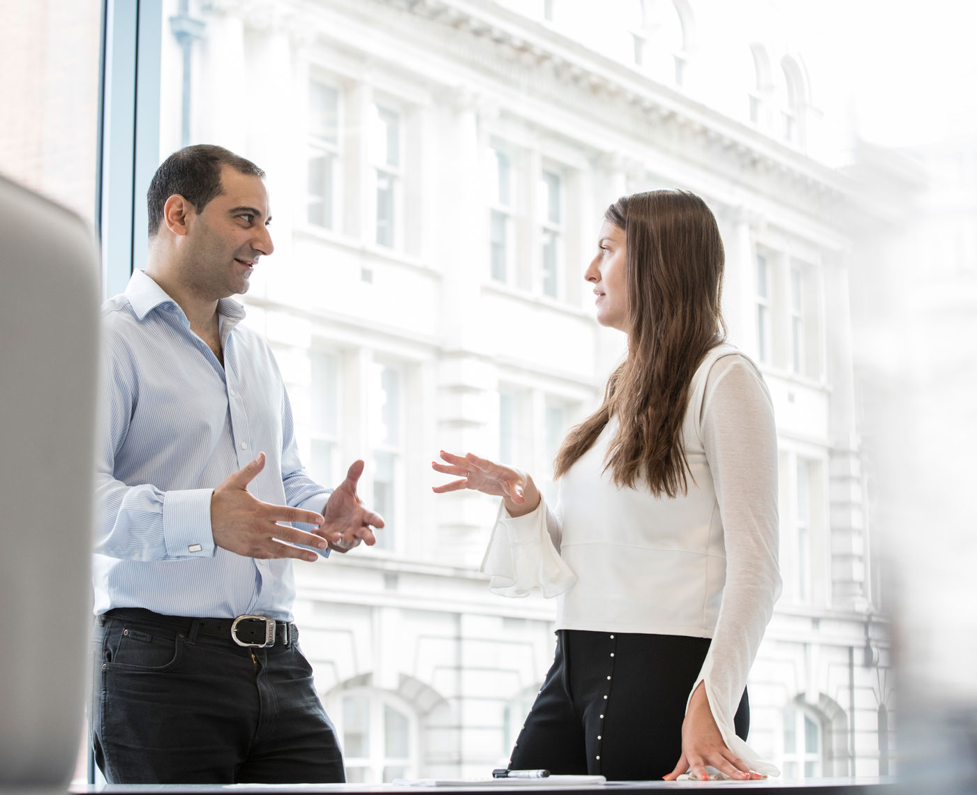 A man and a woman deep in conversation stand by a large window in an office