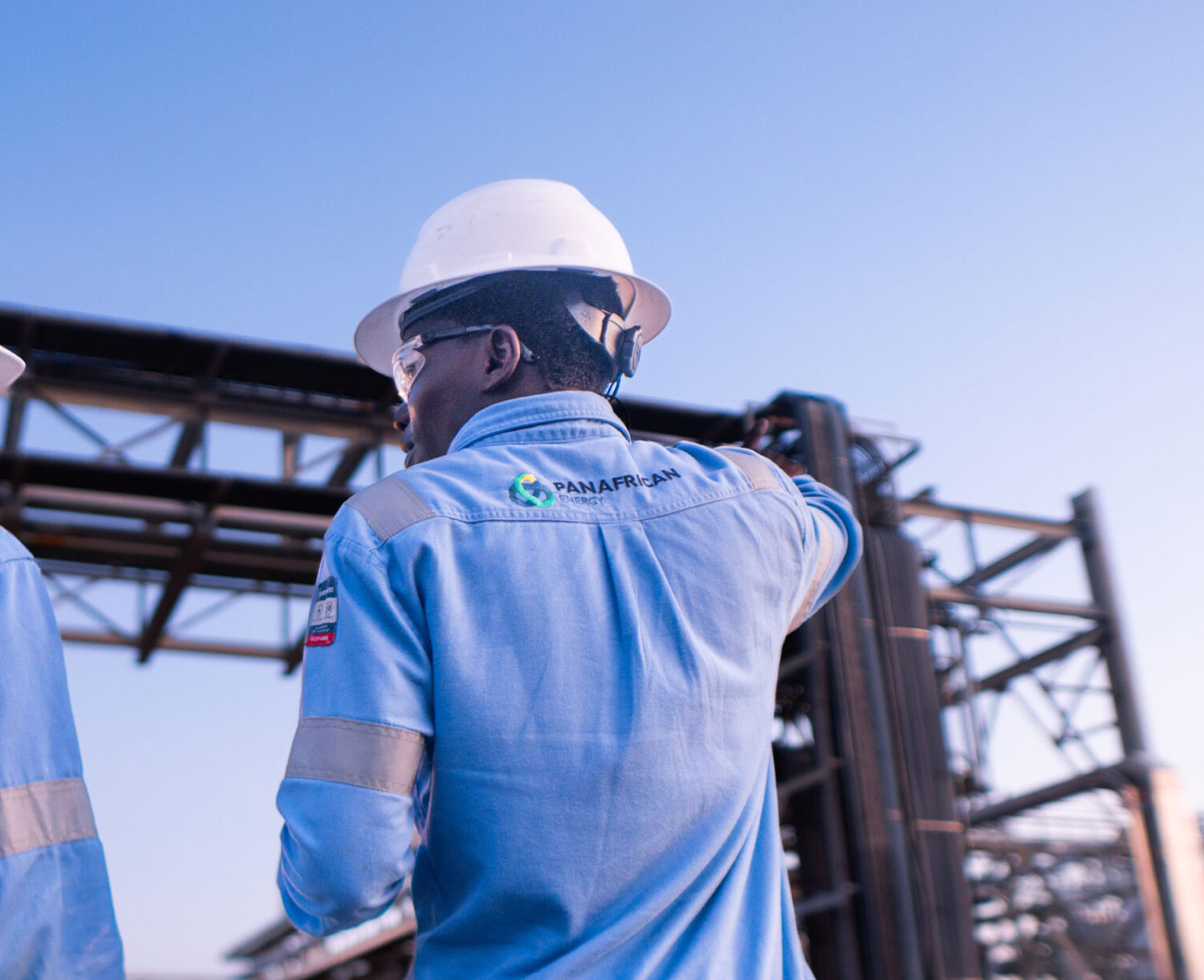 A man wears blue coveralls, a hard hat and safety goggles whilst working outside on a gas plant with the PAET logo on the back of his coveralls