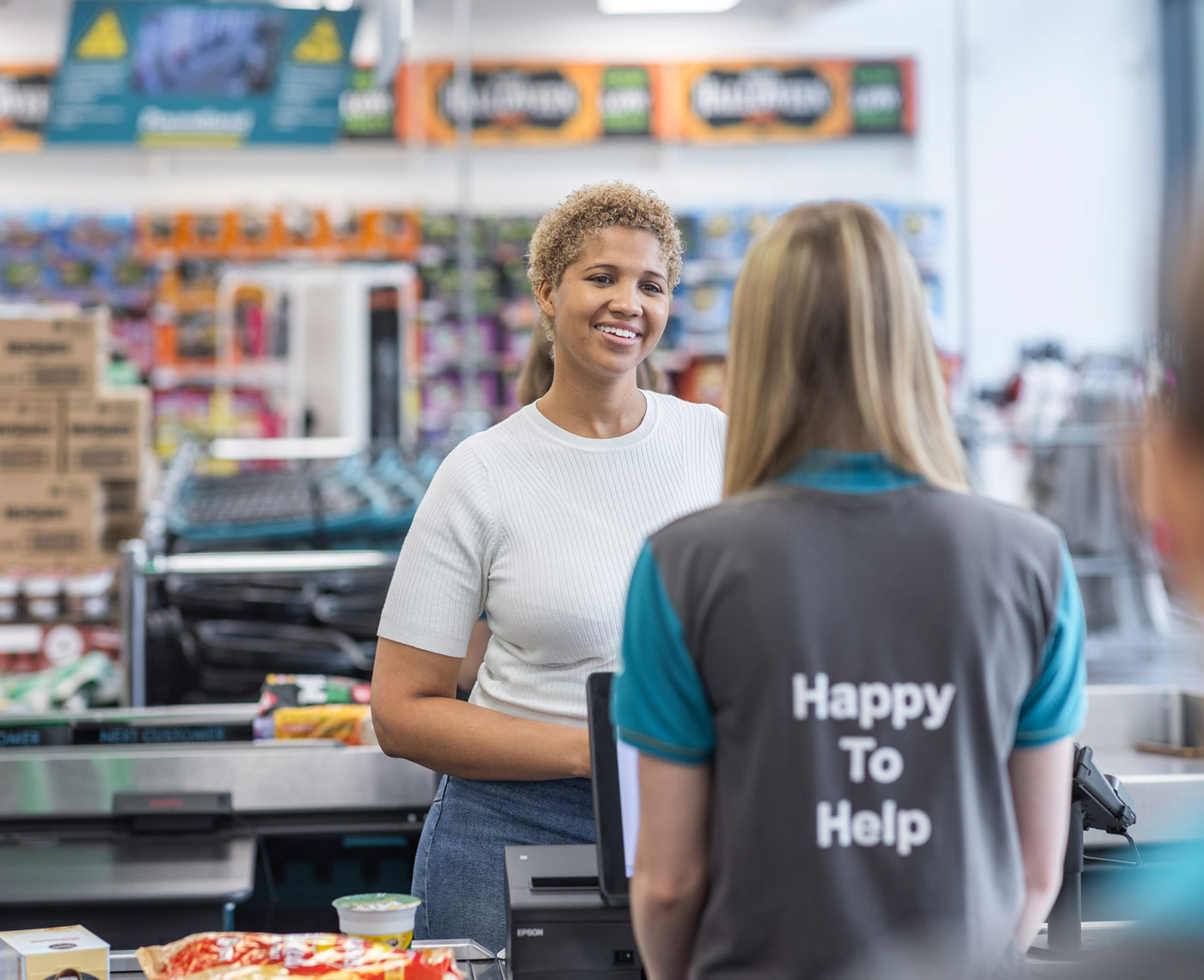 A smiling female customer stands at a checkout talking to a Pepco employee who wears a branded shirt that says: Happy to help